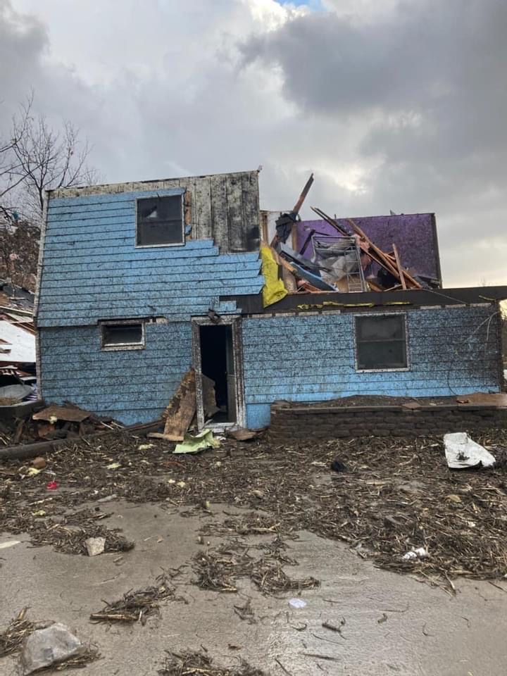 A blue house with a portion of its roof torn off lies on a pile of rubble.