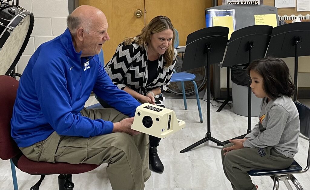 Male and female Lions Club members show the camera they are using to do vision screenings to a child in the screening room.