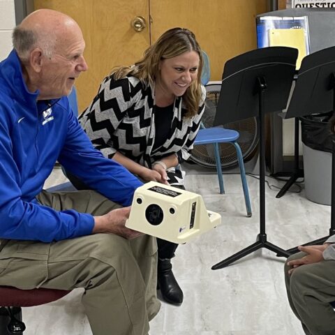 Male and female Lions Club members show the camera they are using to do vision screenings to a child in the screening room.