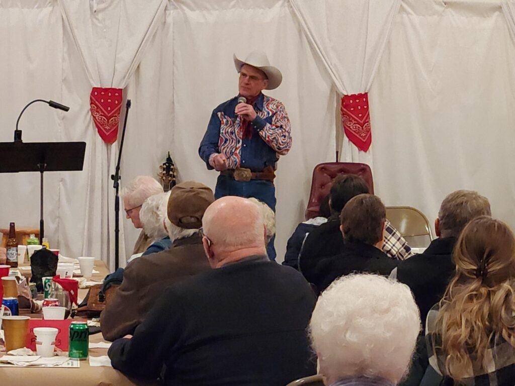 A tall man in a white cowboy hat, western shirt and jeans speaks on a microphone to a room full of people. Behind the man are white curtains tied with red bandanas.