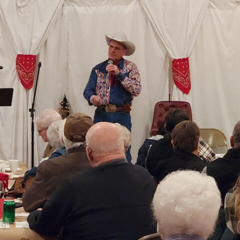 A tall man in a white cowboy hat, western shirt and jeans speaks on a microphone to a room full of people. Behind the man are white curtains tied with red bandanas.